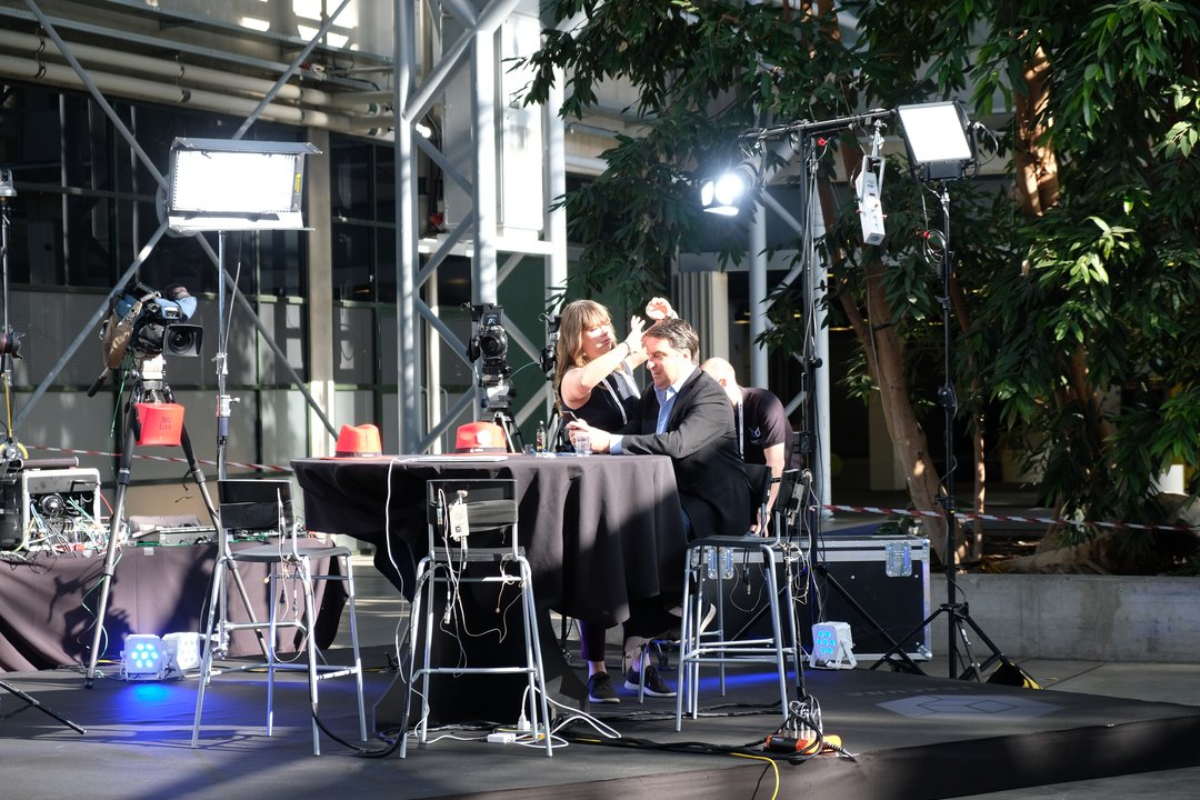 A white male looking at his phone while a hair-dresser prepares him for a video shoot, with plants and audio-video equipment in the background